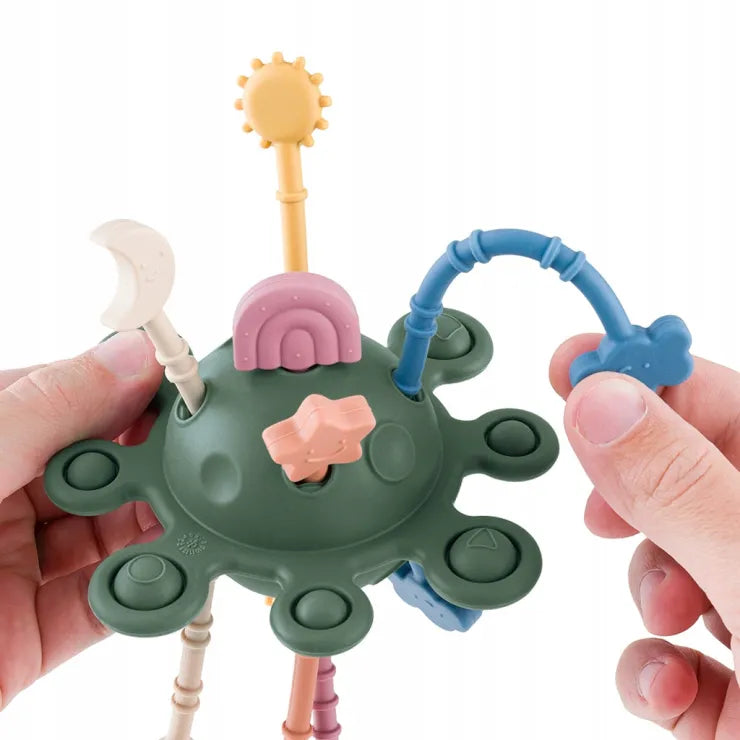 Mãos de adulto a interagir com o brinquedo sensorial "Crazy Planet" verde, mostrando a flexibilidade e os anexos de puxar, como o arco azul.
