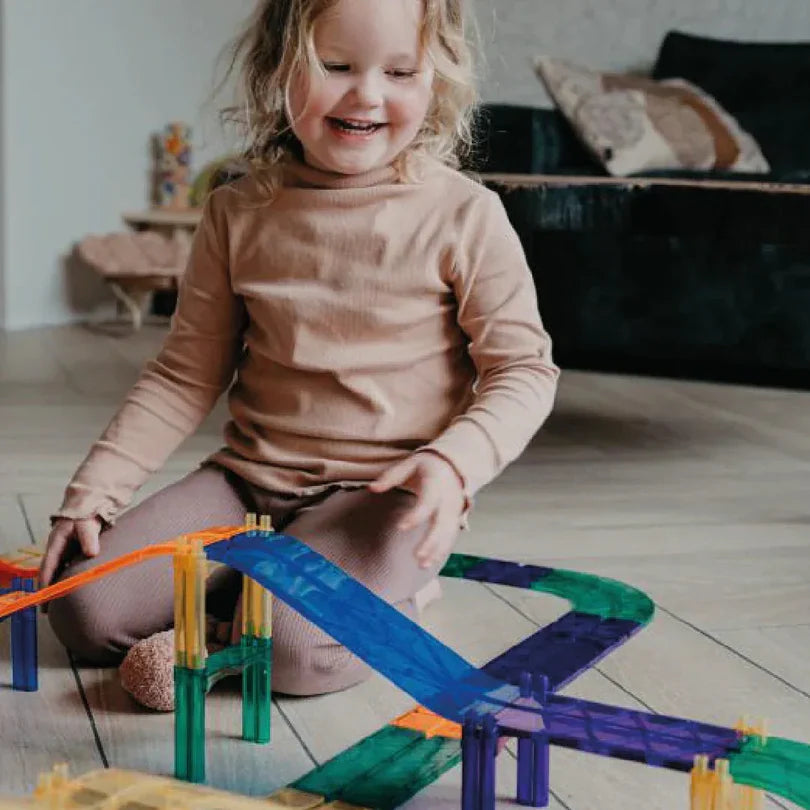 Criança sorridente a brincar no chão com a pista de corrida magnética Cleverclixx em tons pastel, demonstrando envolvimento ativo com o brinquedo.