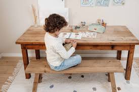 Criança sentada num banco, a brincar com o livro de autocolantes Little Dutch numa mesa de madeira, num ambiente luminoso.