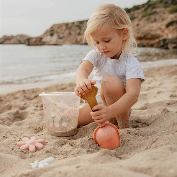 Criança loira a brincar na areia com o balde transparente Little Dutch e acessórios de praia, concentrada a usar uma pequena pá.