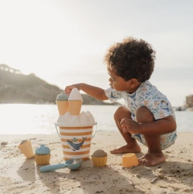 Criança pequena com cabelo encaracolado a brincar na praia com o conjunto de gelados de areia Little Dutch, colocando um topo de gelado no balde listrado.