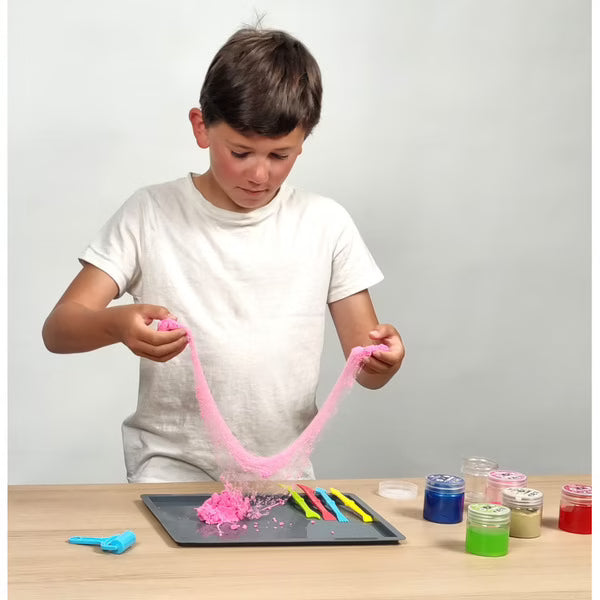 Criança a manipular um slime rosa, esticando-o sobre um tapete de brincar, com vários potes de slime ao lado.