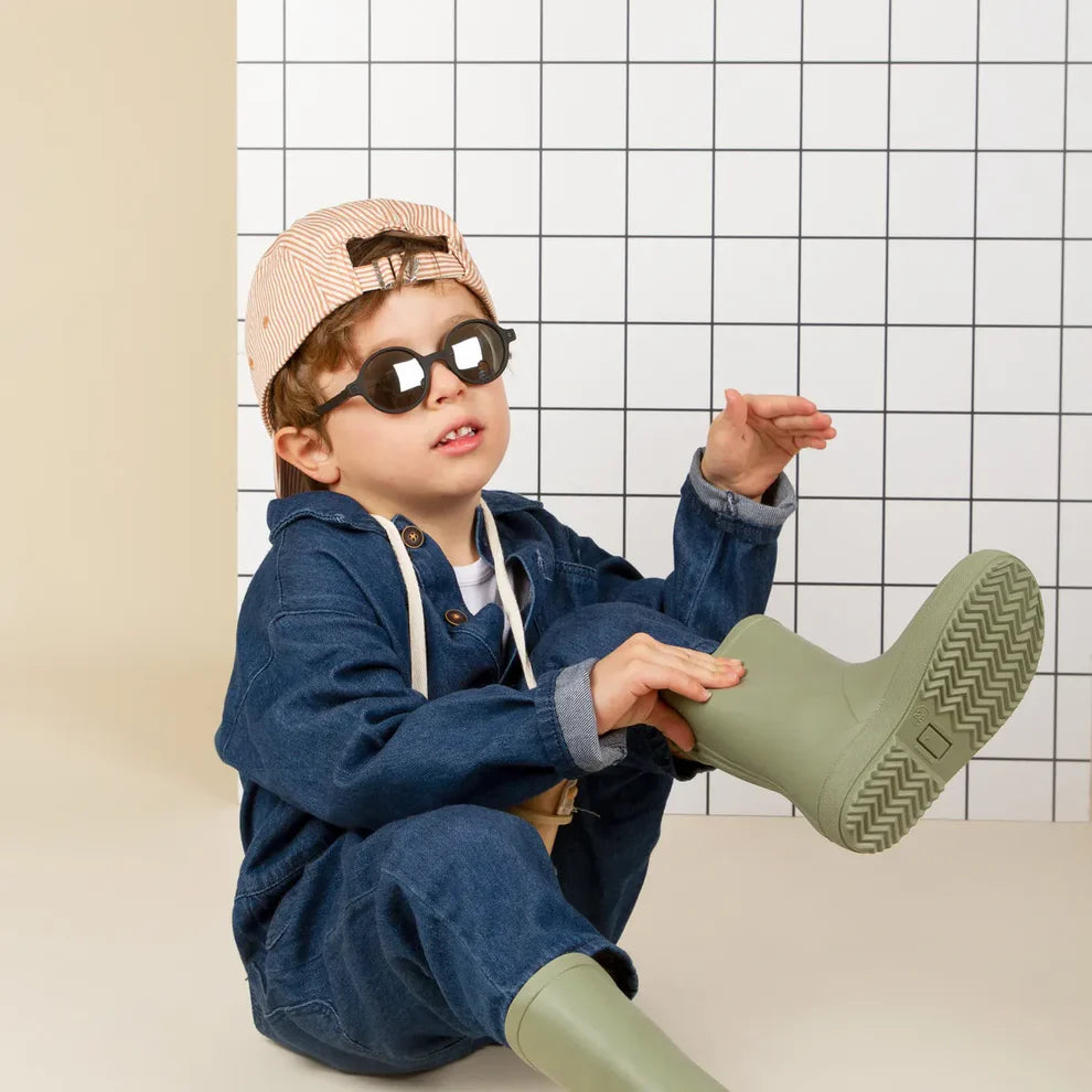 Criança com boné e óculos de sol Kietla ROZZ pretos, a calçar galochas verdes, num cenário com fundo de azulejos brancos e pretos.