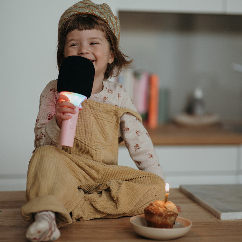 Criança sorridente com gorro a cantar no microfone de karaoke KidyWolf com luzes, sentada perto de um muffin com vela.