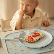 Bebé sorridente sentado à mesa a comer uma fatia de pão com morangos sobre o prato do conjunto Mepal Mio, utilizando a faca infantil.