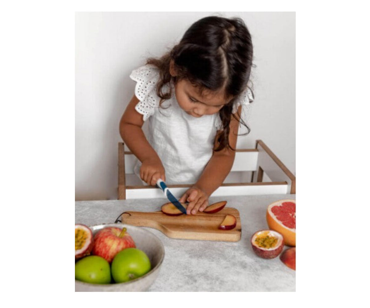 Criança pequena a usar uma faca KiddiKutter azul para cortar fatias de ameixa numa tábua de madeira, promovendo a autonomia na cozinha.