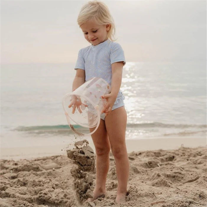 Criança de pé na praia, a despejar areia de um balde transparente Little Dutch para construir uma estrutura na areia.