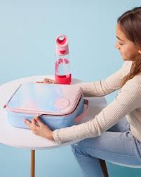 Criança a fechar uma lancheira b.box com padrão de nuvens, ao lado de uma garrafa de água empilhável cor-de-rosa e vermelha, sobre uma mesa branca.