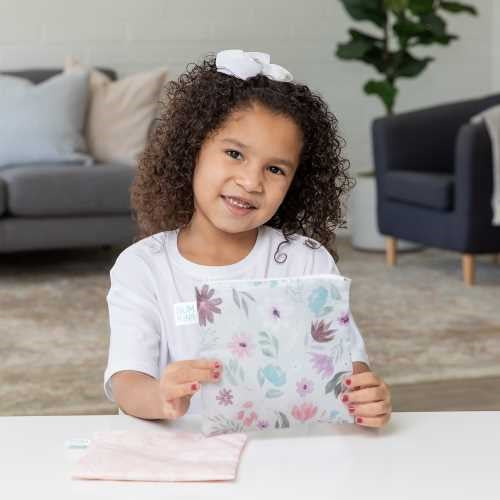 Criança sorridente com cabelo encaracolado a segurar um dos sacos reutilizáveis florais Bumkins, demonstrando o uso prático do produto.