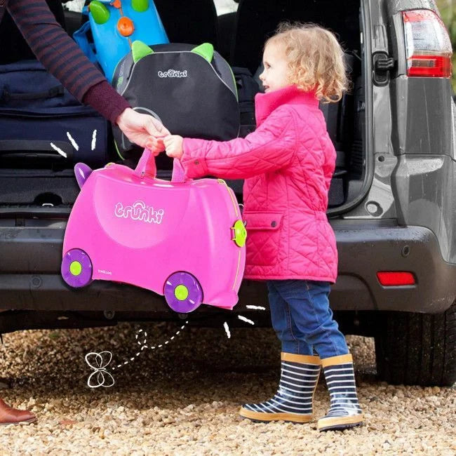 Criança de casaco rosa a receber uma mala Trunki rosa de um adulto junto à traseira de um carro cinzento.
