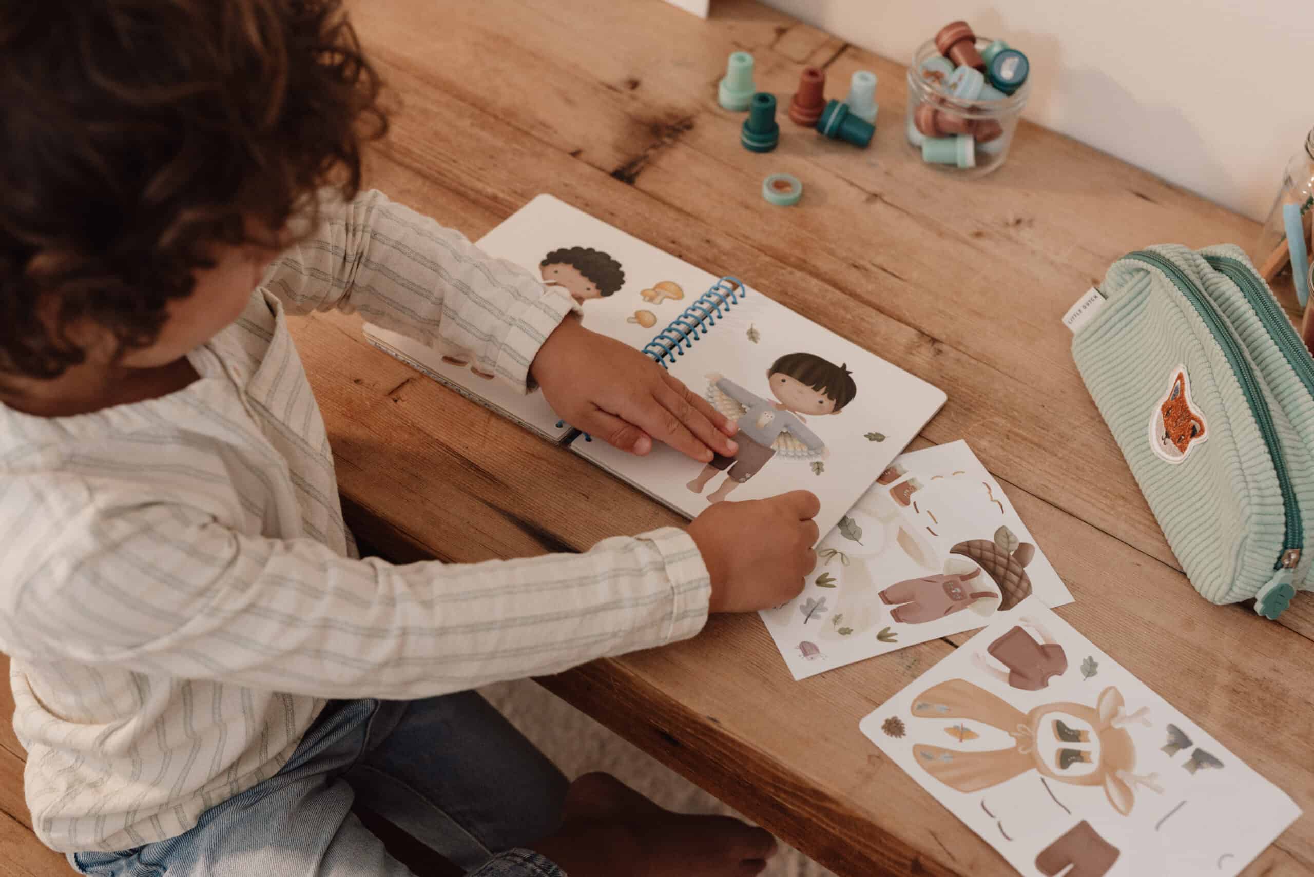 Criança a utilizar o livro de autocolantes "Dress Me Up" sobre uma mesa de madeira, aplicando autocolantes no livro.