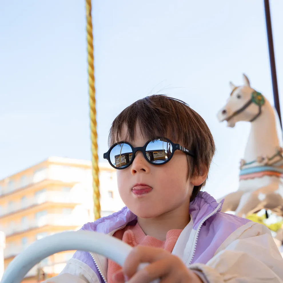Criança a usar os óculos de sol Kietla ROZZ pretos enquanto conduz um carrossel, com a língua de fora.