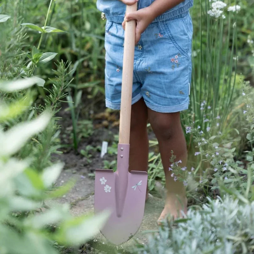 Criança a segurar a pá de jardinagem cor-de-rosa Little Dutch, com as mãos no cabo de madeira, em pé num jardim com vegetação densa.