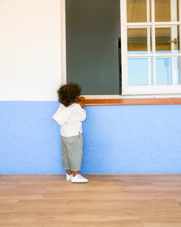 Criança de costas, a usar os ténis Muris Petranova, a olhar pela janela aberta de uma casa com parede azul.