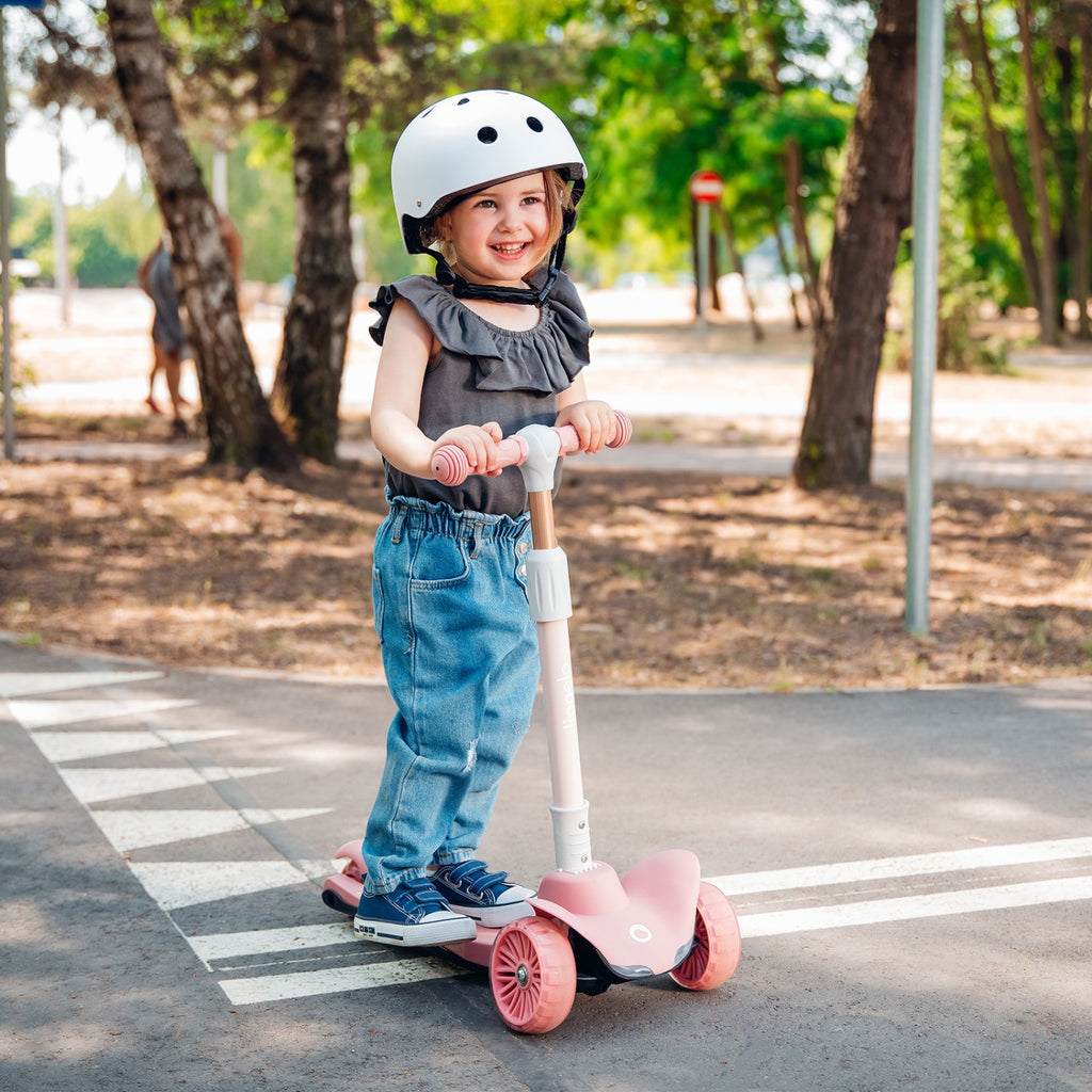 Criança sorridente a usar a trotinete Lionelo Timmy rosa com capacete branco, a andar num passeio com marcações de peões.