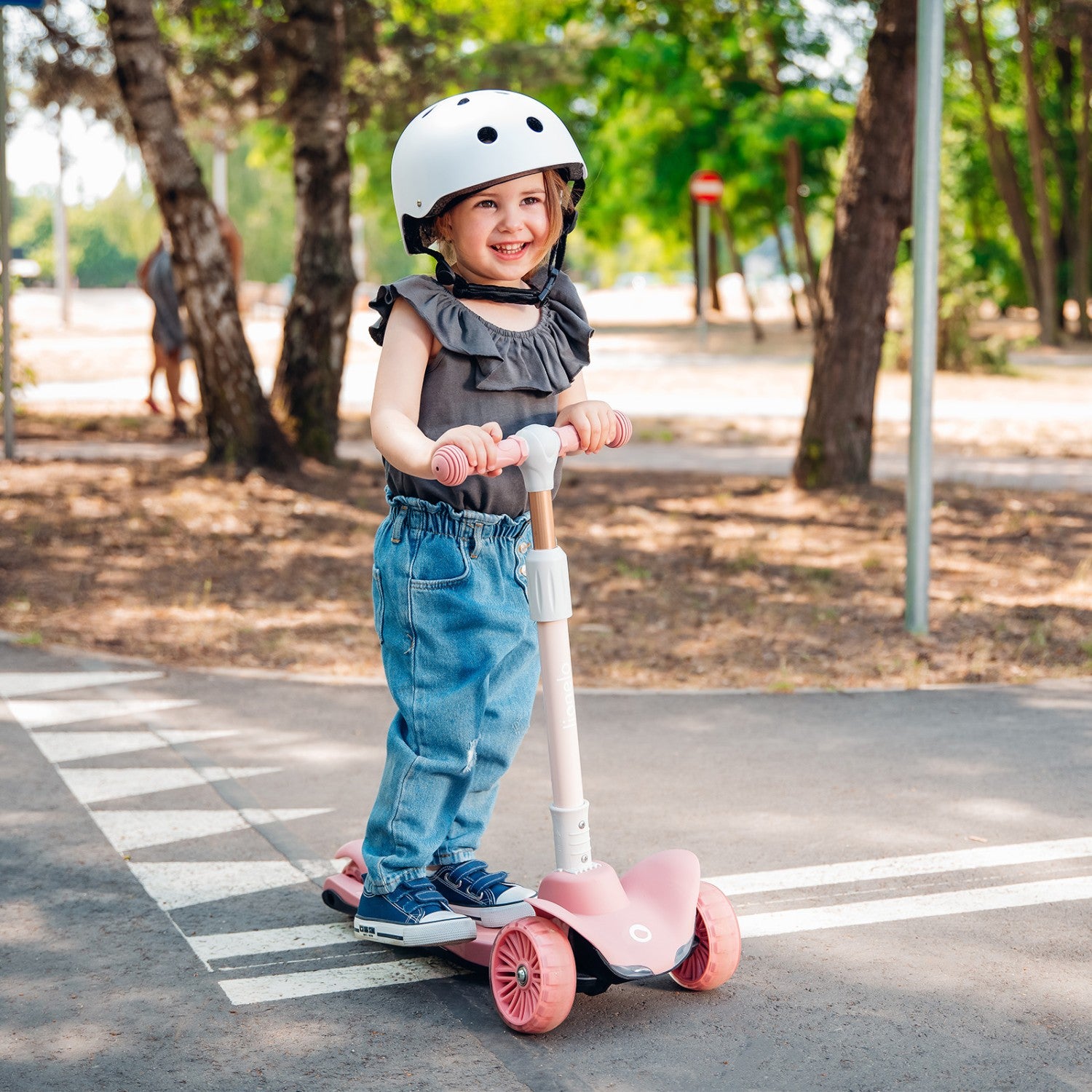 Criança sorridente a usar a trotinete Lionelo Timmy rosa com capacete branco, a andar num passeio com marcações de peões.