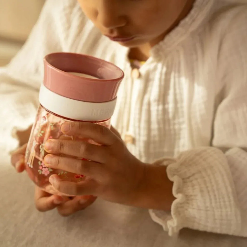 Criança pequena a segurar e a observar o copo de treino Little Dutch Mepal cor de rosa com padrão floral.
