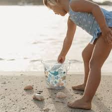 Criança de fato de banho azul a apanhar areia com a mão para colocar no balde decorado na praia.