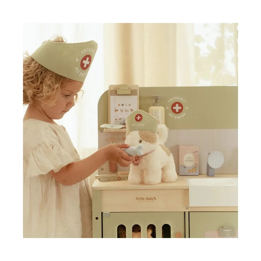 Criança pequena vestida com gorro de veterinário a examinar um peluche de cão com um instrumento médico na sua clínica de brincar.