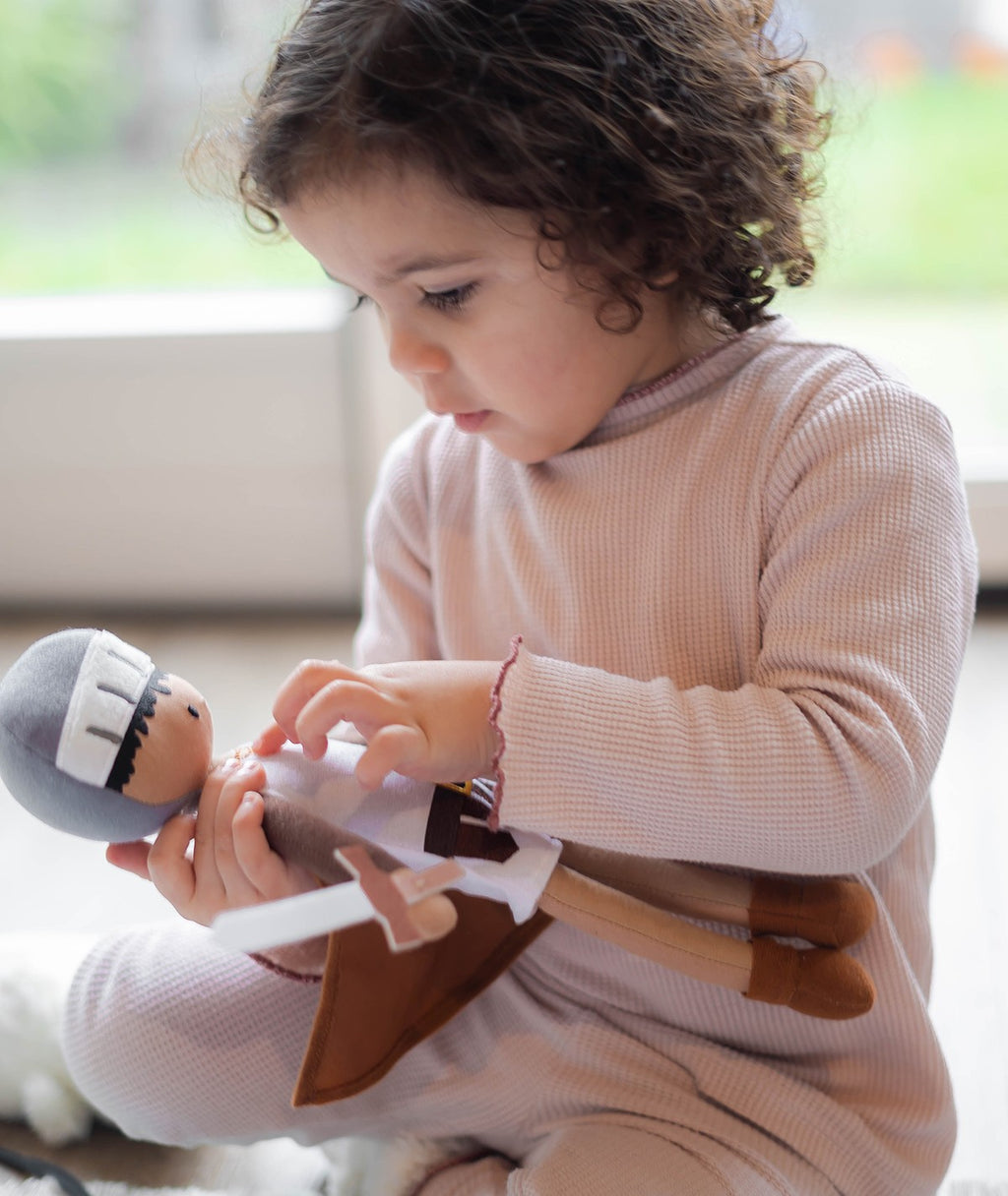 Criança pequena com pijama cor-de-rosa a brincar com o peluche Cavaleiro Kai, examinando os detalhes do boneco.