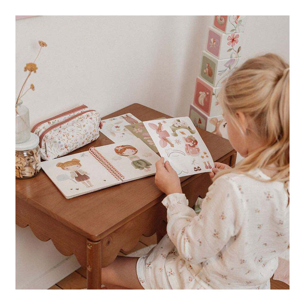 Criança loira sentada a brincar com o livro de vestir Little Dutch sobre uma mesa de madeira, manuseando as páginas e autocolantes.