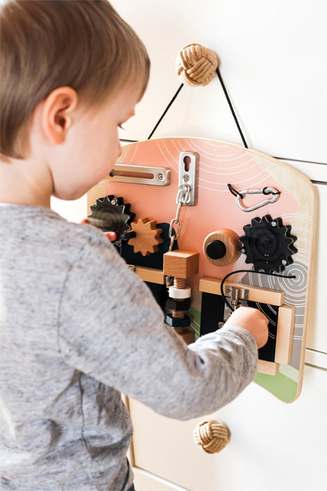 Criança a brincar com o quadro de atividades small foot pendurado numa parede, mostrando a praticidade da alça e a versatilidade do brinquedo.