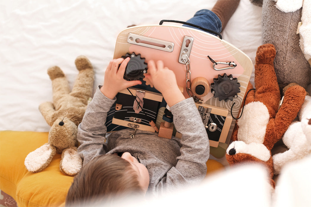 Vista aérea de uma criança a brincar com o quadro de atividades small foot, rodeada de brinquedos de pelúcia, mostrando o contexto lúdico e aconchegante do produto.
