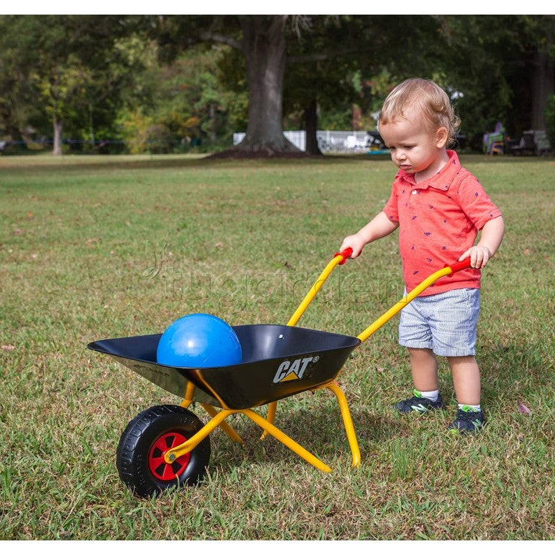 Criança pequena a brincar num parque com o carrinho de mão infantil Rolly Metal CAT, transportando uma bola azul, num ambiente verde e natural.