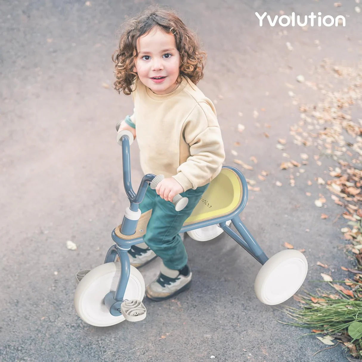 Criança pequena a pedalar alegremente o triciclo Yvolution Classic Trike azul num caminho de jardim.