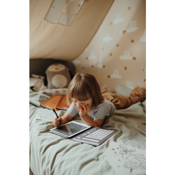Criança pequena deitada numa cama, concentrada a desenhar no tablet LCD KidyWolf com a caneta, num ambiente acolhedor de tenda.