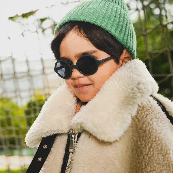 Detalhe de uma criança a usar óculos de sol pretos Kietla, com gorro verde e casaco de pelo, focando no rosto.