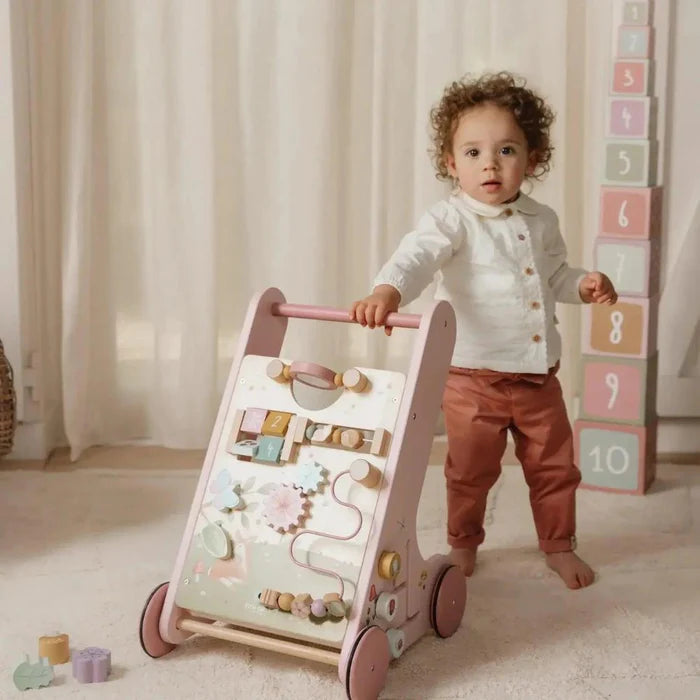 Criança em pé ao lado do andarilho de atividades Little Dutch, com um painel de medição de altura visível ao fundo.