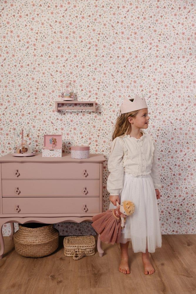 Criança com coroa de feltro a segurar a Boneca Princesa Julia junto a uma cómoda rosa, num quarto decorado com tema primaveril.