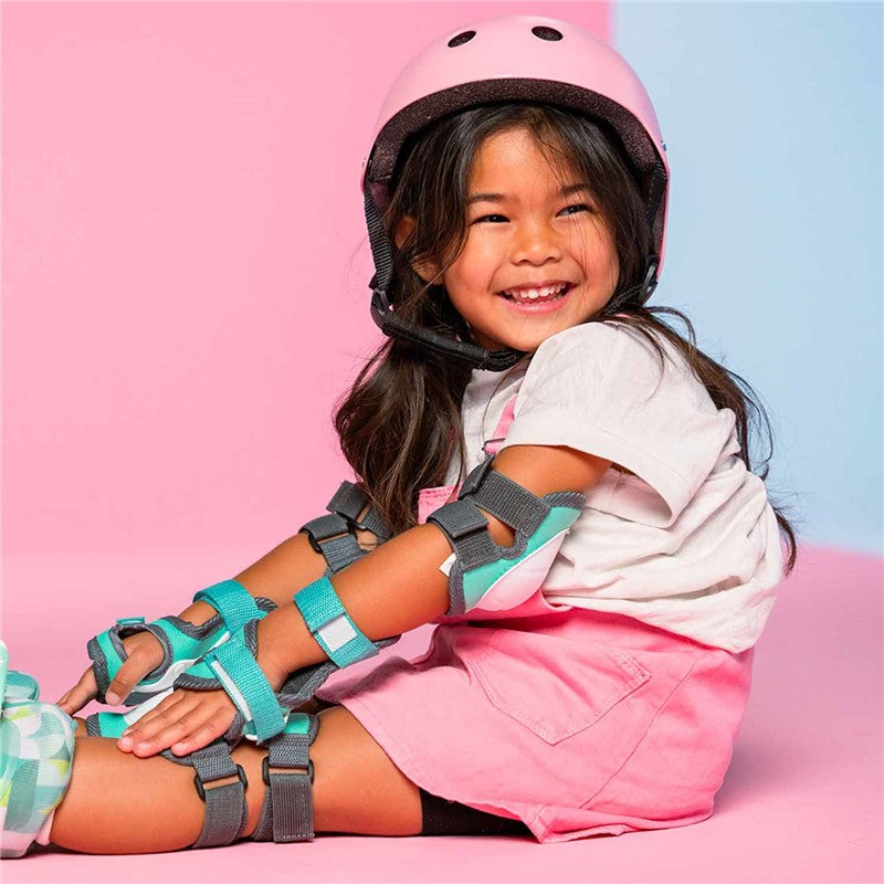 Criança sorridente a usar um capacete rosa e um conjunto de proteções Yvolution turquesa e cinzento, sentada num fundo colorido.