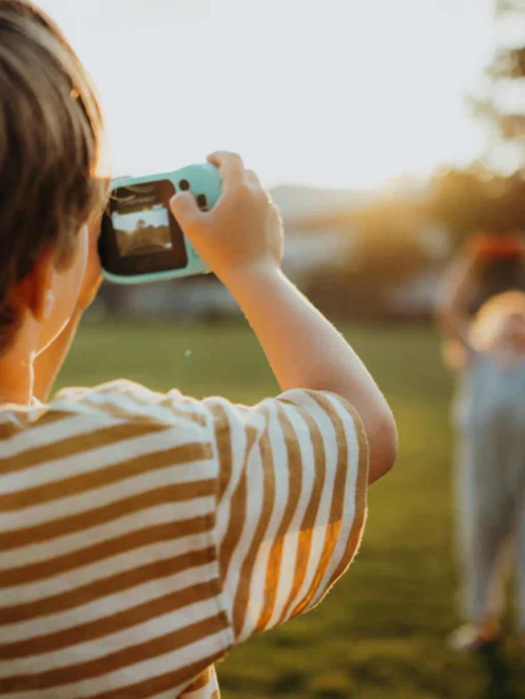 Criança de costas, a usar a máquina Kidywolf azul-clara para fotografar alguém ao pôr do sol num relvado, focando na ação de fotografar.