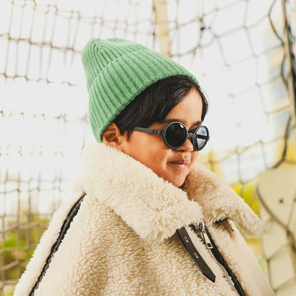 Criança a usar gorro verde e óculos de sol pretos Kietla, sorrindo, com um casaco de pelo sintético.