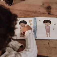 Vista de cima, desfocada, de uma criança a interagir com o livro de autocolantes "Dress Me Up" na mesa.