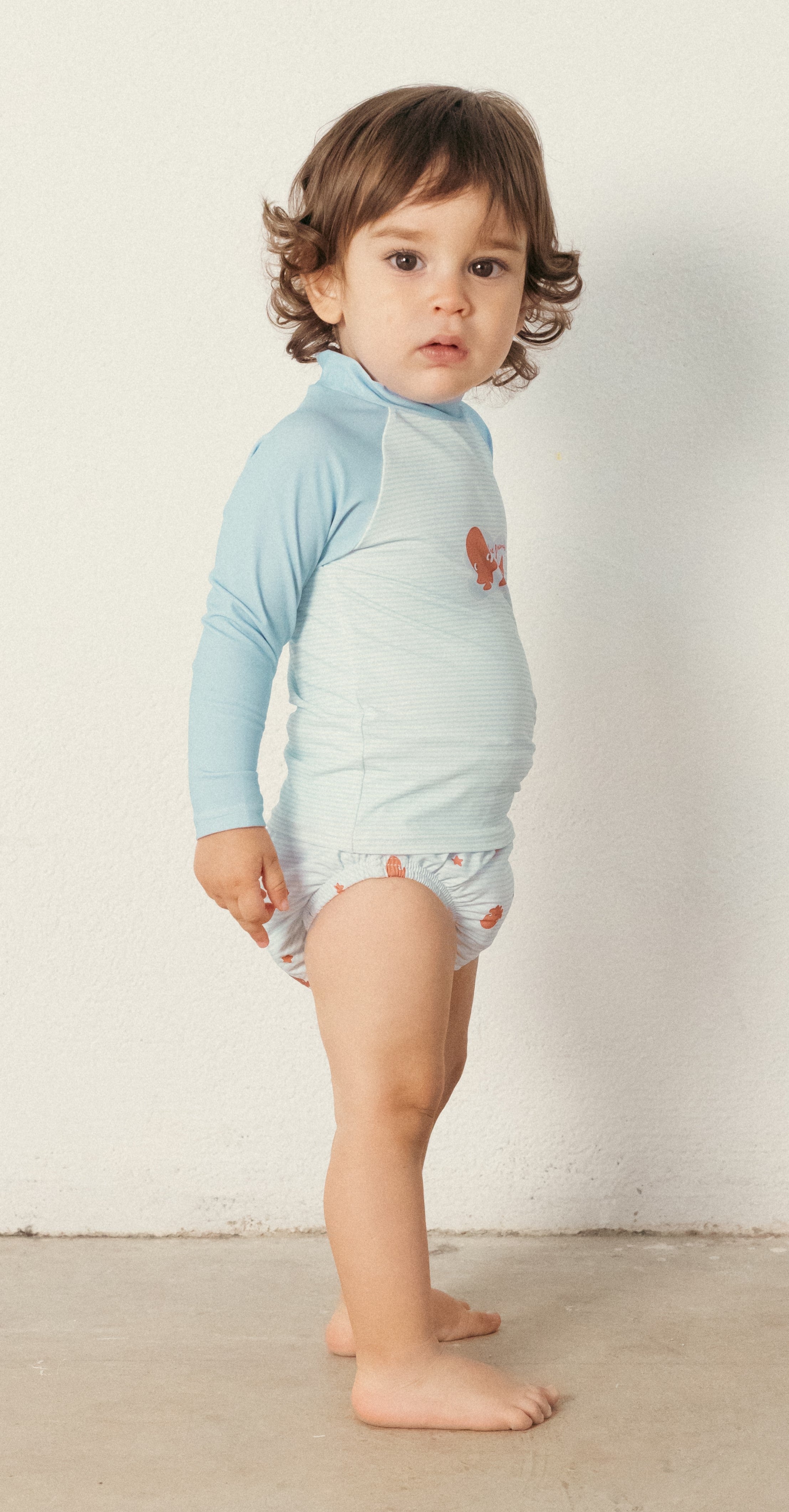Criança pequena em pé, vestindo a cueca de praia Waterlemon com padrão de polvos e camisola de banho azul de manga comprida.