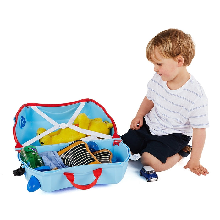 Criança pequena sentada ao lado da sua mala Trunki azul aberta, a organizar brinquedos e roupa no interior.