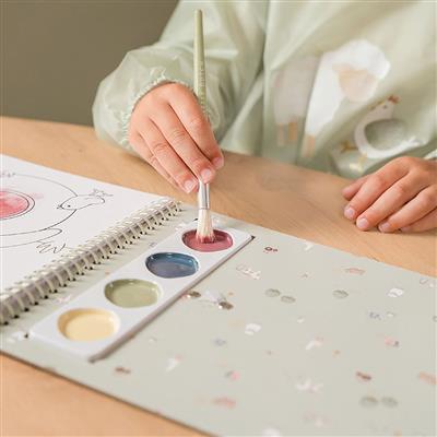 Criança a usar o pincel para aplicar aguarela numa das páginas ilustradas do livro, com a paleta de cores em primeiro plano.