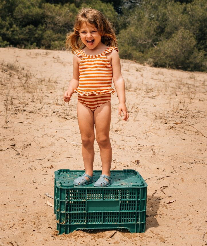 Criança de pé numa caixa verde na areia, vestindo um fato de banho e segurando as sandálias Igor Barefoot. Cenário de verão, ideal para praia e diversão ao ar livre.