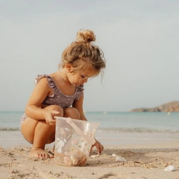 Criança agachada na areia, a recolher conchas e a colocá-las no balde de praia transparente Little Dutch, com o mar ao fundo.