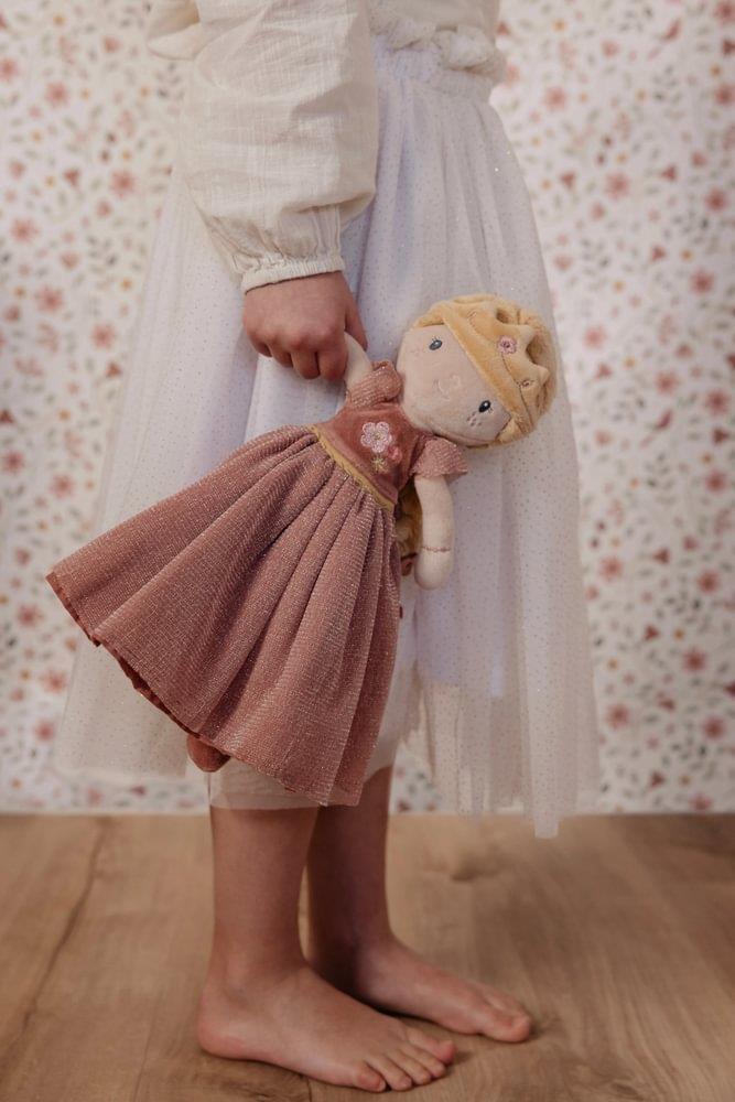 Criança a segurar a Boneca Princesa Julia pela mão, enquanto veste um vestido branco com brilho, sobre fundo de papel de parede floral.