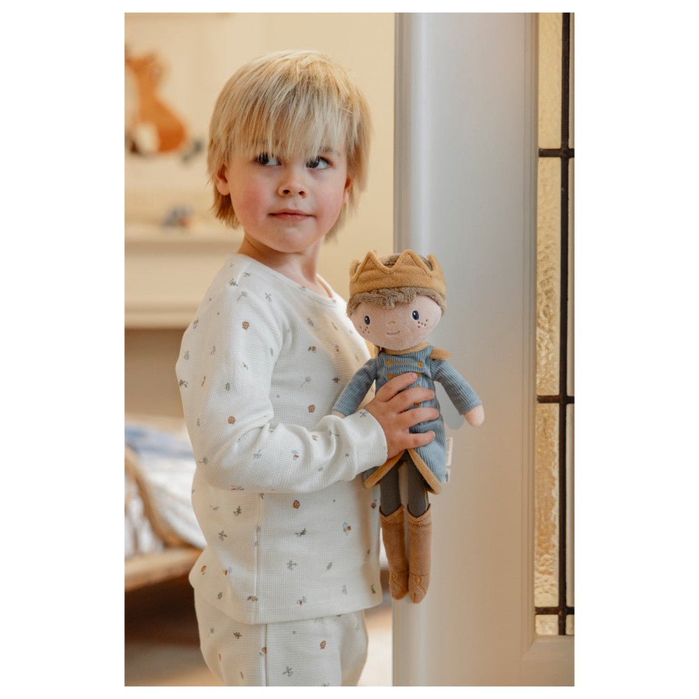 Criança loira de pijama branco a segurar o boneco Príncipe Jim da Little Dutch junto a uma porta.