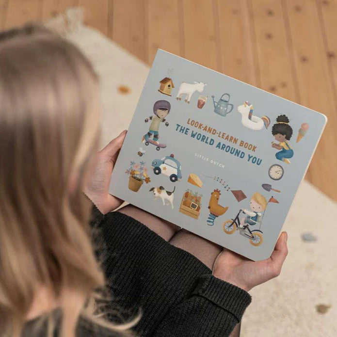 Criança a segurar o livro ilustrado "Look-and-Learn Book The World Around You" da Little Dutch, mostrando a capa com várias ilustrações coloridas.