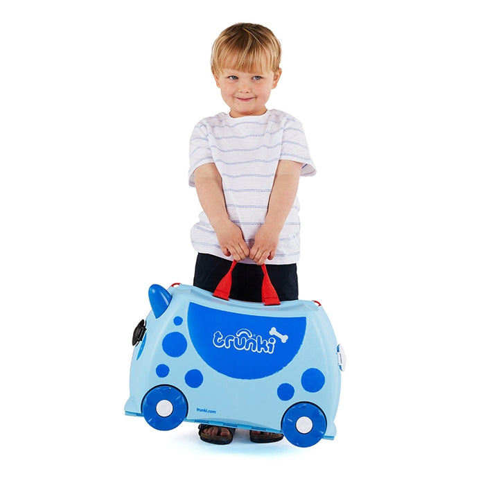Criança loira sorridente a segurar as pegas vermelhas da sua mala Trunki azul, pronta para viajar.