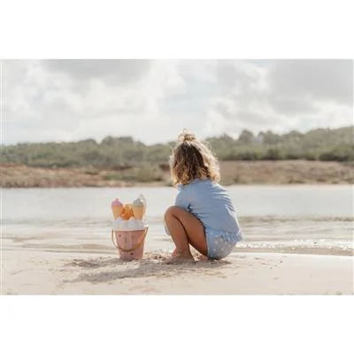 Criança pequena sentada de costas na areia, a brincar com o balde de gelados de areia Little Dutch junto à água.