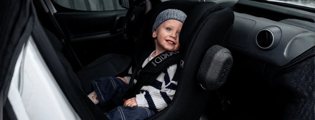 Criança sorridente, com gorro, instalada na cadeira auto AXKID Minikid CORE no banco traseiro de um veículo.