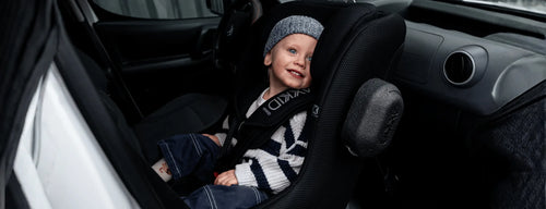 Criança sorridente, com gorro, instalada na cadeira auto AXKID Minikid CORE no banco traseiro de um veículo.