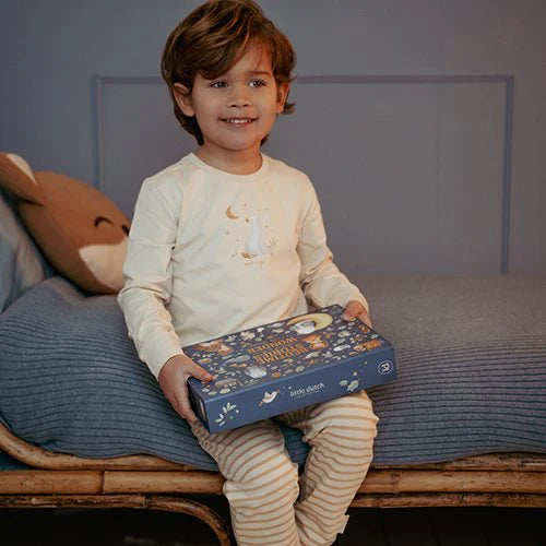 Criança sentada na cama, sorridente, a segurar a caixa do projetor lanterna Little Dutch.